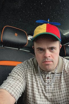 A serious man in a propeller beanie and chain necklace sitting in the back seat of a luxury car at night
