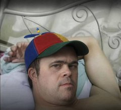 A man in a propeller beanie lying in bed, staring past the camera with a heavy expression