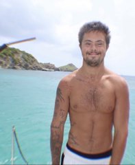 A shirtless man smiling on a boat in turquoise water, a propeller blade visible in the corner
