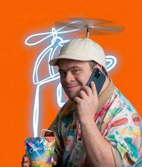 A smiling man on the phone holding a Capri Sun, in front of an orange wall with a white crayon doodle of a propeller beanie