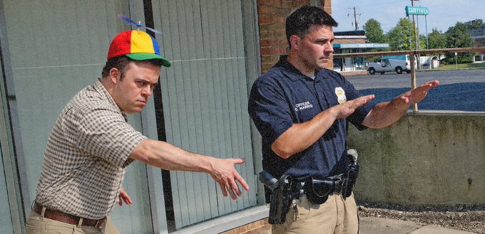 A police officer holding both hands up to stop a man in a propeller beanie who is reaching forward dramatically