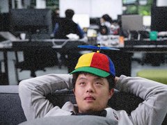 A man in a propeller beanie leaning back with hands behind his head in a dark open-plan office