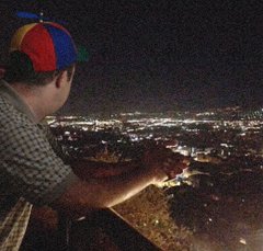 A man in a propeller beanie looking out over a glittering city at night from a high overlook