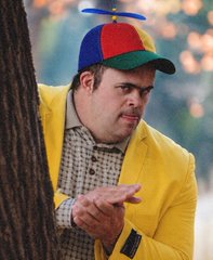 A man in a propeller beanie and bright yellow blazer peeking out from behind a tree, rubbing his hands