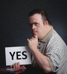 A man holding up a white card that says YES in large black letters, finger on his chin